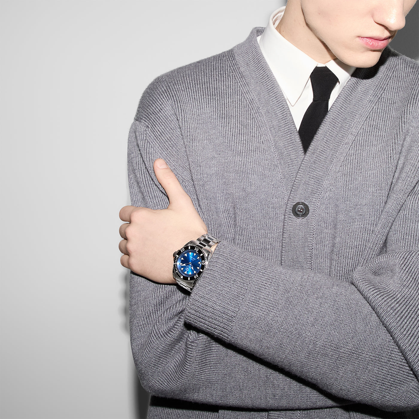 A person crosses their arms while wearing a gray cardigan over a white shirt and black tie, showcasing a Gucci Dive Automatic 40mm Watch from GUCCI on their left wrist. The steel case of this timepiece with its blue dial stands out against the plain, light-colored background.