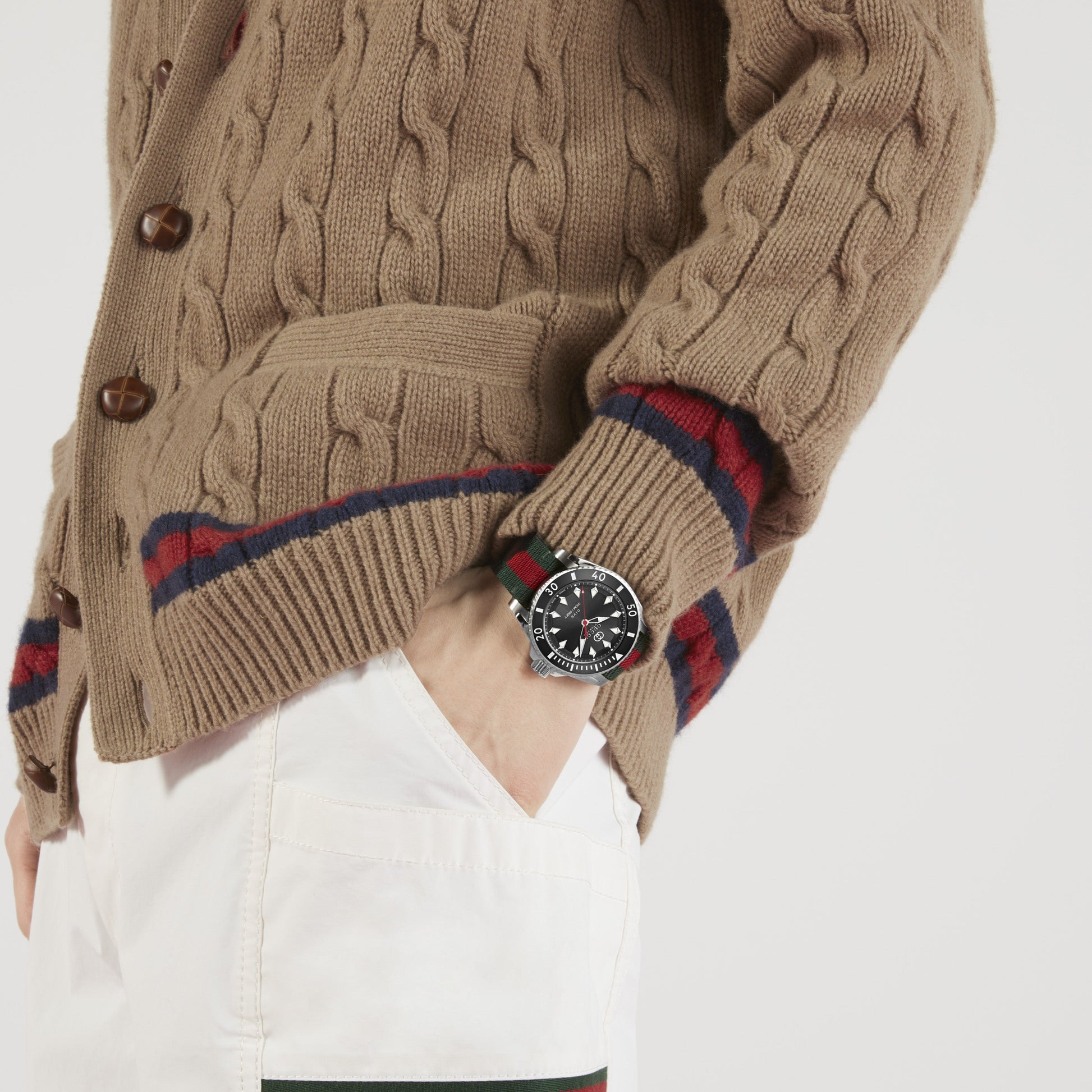 A person is wearing a beige cable-knit sweater and white pants, with a GUCCI Watch featuring the Interlocking G logo and a black and red striped band. Their hand is in the pocket of the pants, prominently displaying the Gucci Dive Automatic 40mm Watch's face, exuding a bold style characteristic of Gucci Dive collections.