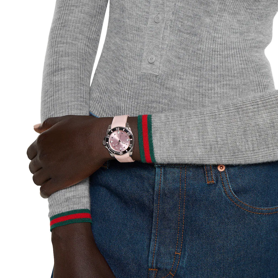 A person in a gray long-sleeve henley with green and red striped cuffs and blue jeans crosses their arms at the waist, wearing a GUCCI Watch Gucci Dive Automatic 36mm with a pink rubber strap on their left wrist.