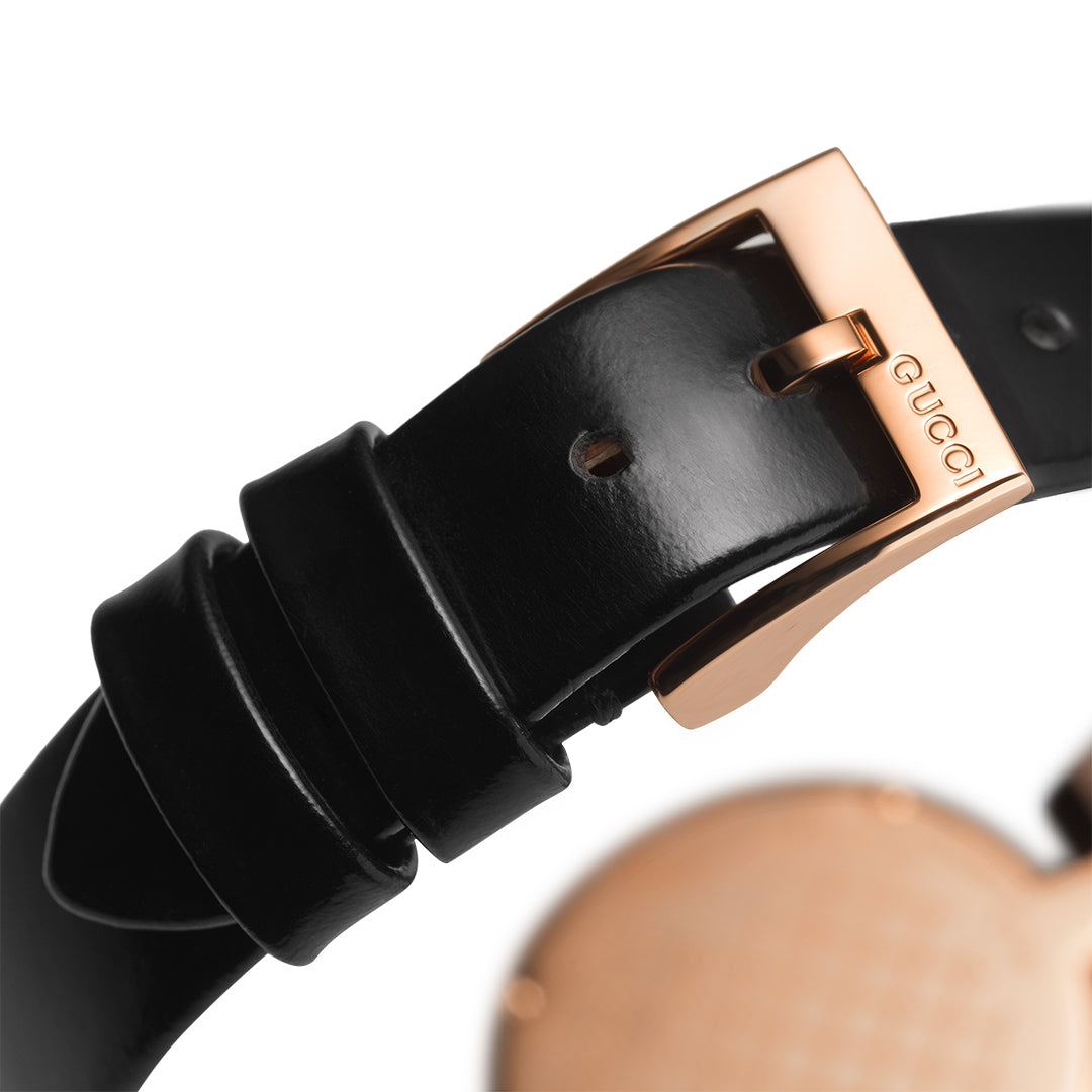 A detailed view of the black leather strap featuring a gold buckle, belonging to the Gucci Diamantissima Quartz 27mm Watch collection. The strap includes adjustment holes, while their signature elegantly etched on the rose gold case shines in the backdrop next to a softly focused watch face.