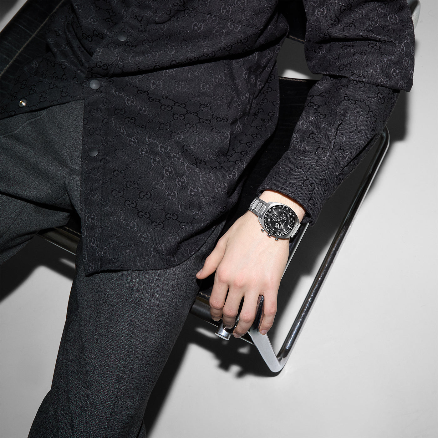 A person sits on a chair wearing a black patterned shirt, gray pants, and a GUCCI Watch Interlocking Quartz 41mm with stainless steel bracelet. Only the torso and legs are visible, with their arm resting casually on the chair's edge. The setting is minimalist and modern.