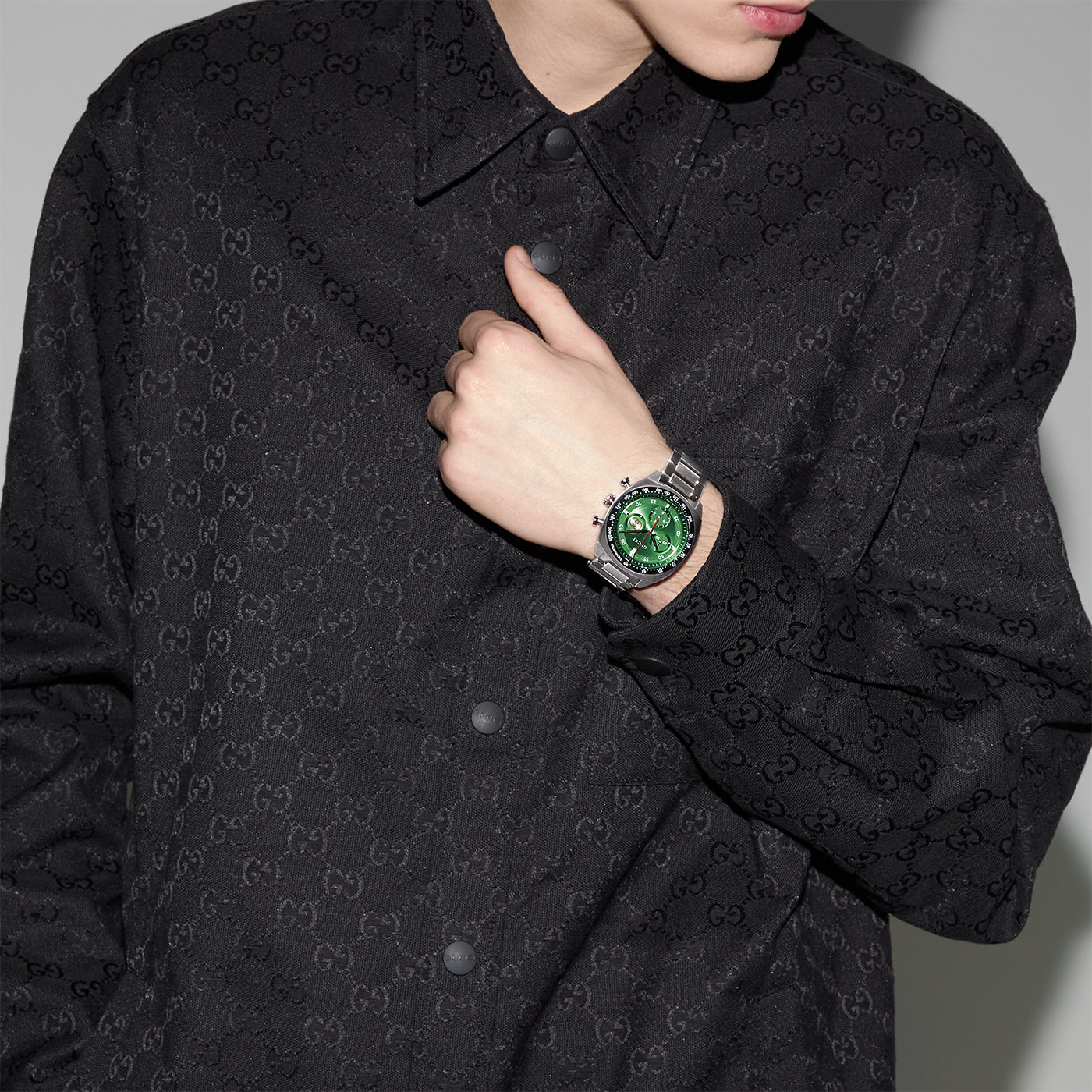 A person wearing a black Gucci jacket with a subtle logo pattern is posing with their hand on their chest, showcasing the Gucci Interlocking Quartz 41mm Watch by GUCCI, featuring a stainless steel bracelet and a bright green face.