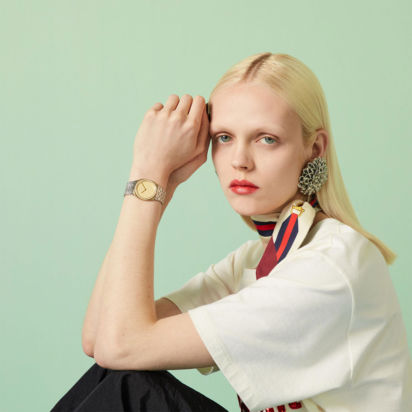 A person with long blonde hair sits against a light green background, wearing a white shirt, large floral earrings, and a striped scarf. As they rest their chin on their hand, they showcase the luxurious Gucci 25H Quartz 30mm Watch from GUCCI on their wrist.