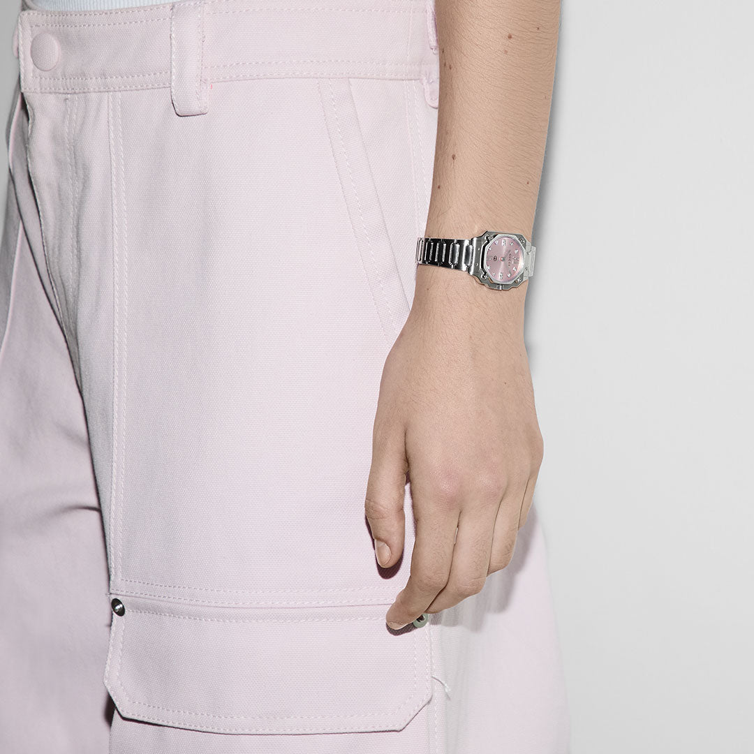 A person in pale pink pants with large pockets wears a GUCCI G-Flat Quartz 24mm watch with a pink face and diamond indexes on their left wrist, against a plain light-colored wall.