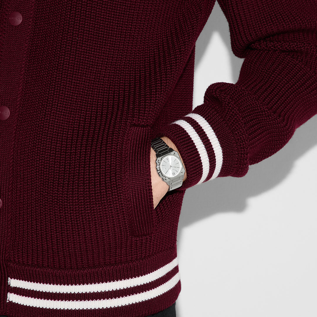 A person in a maroon knit sweater with white striped cuffs and waistband has their hand in their pocket, displaying a GUCCI Watch G-Flat Quartz 30mm with a silver galvanized dial and stainless steel band.