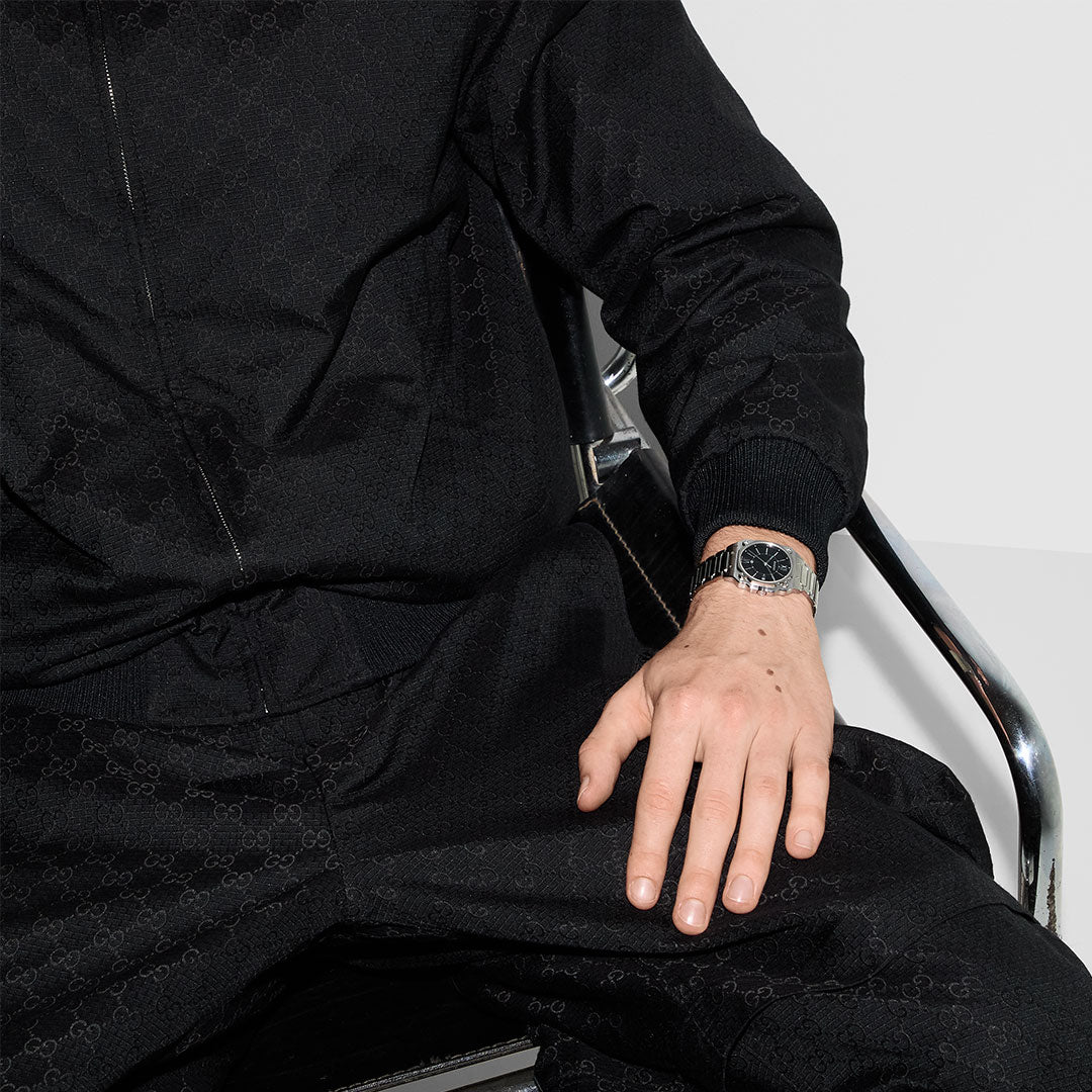 A person in a black patterned tracksuit sits on a chrome chair, their left hand on their leg, wearing a GUCCI G-Flat Quartz 30mm watch with a black galvanized dial. The scene features a white, minimalistic background.