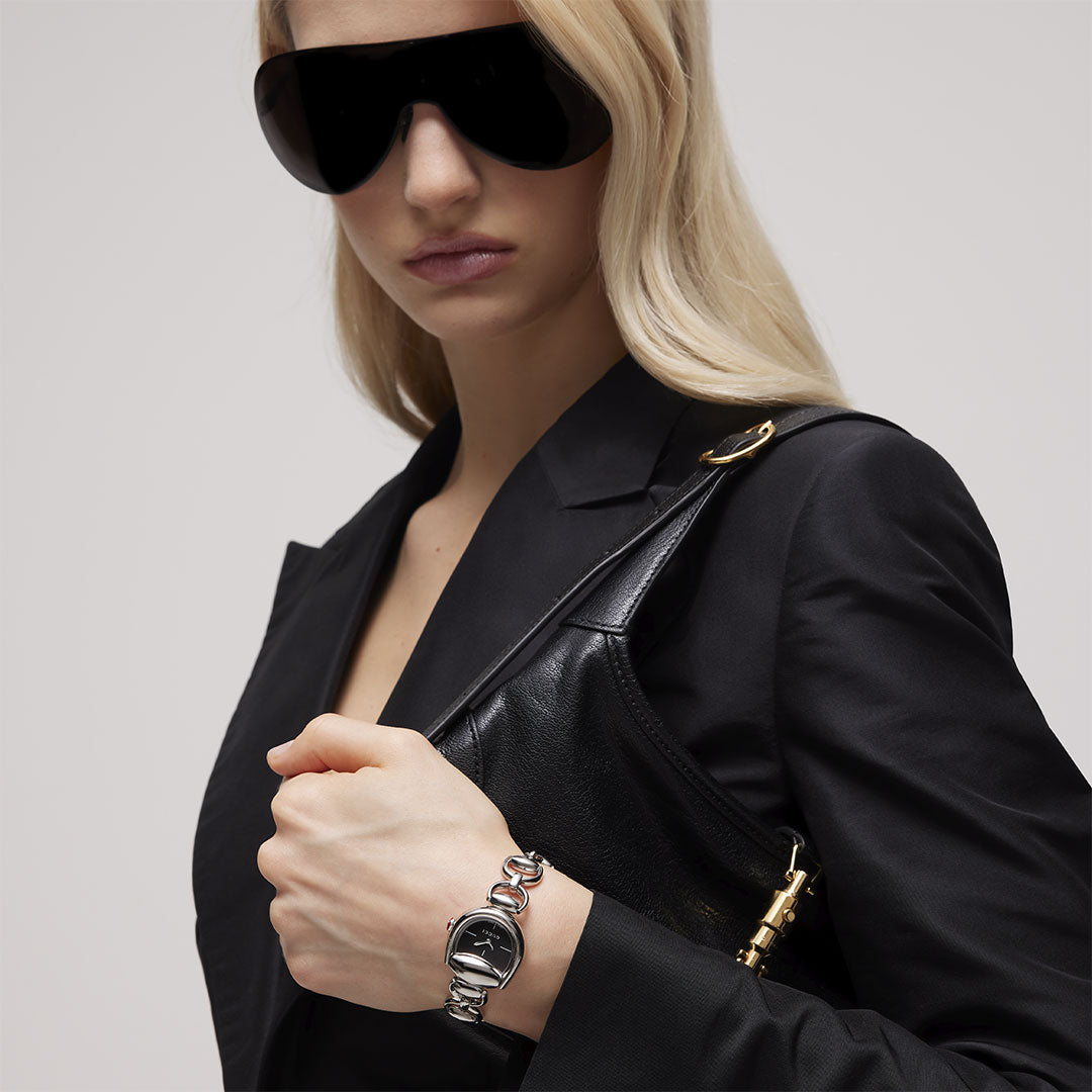 A woman with long blonde hair wears oversized black sunglasses, a black blazer, and holds a black handbag. She accessorizes with a GUCCI Watch—specifically the Gucci Horsebit Quartz 27mm x 25mm—and a chunky silver and black bracelet.