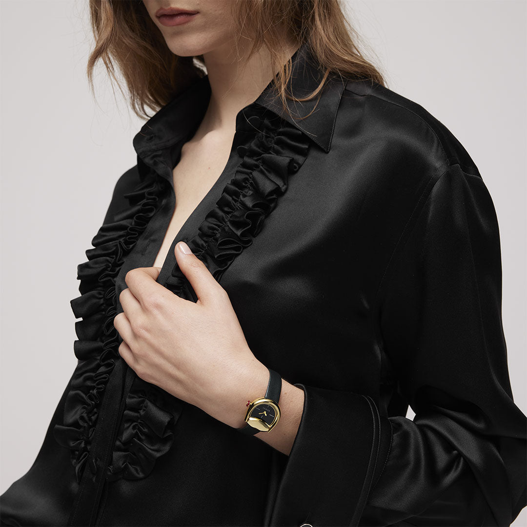 A woman in a black satin blouse with ruffled trim wears a GUCCI Watch Horsebit Quartz 27mm x 25mm with a yellow gold-plated case, holding her left hand to her chest. Her loose hair and face are partially out of frame.