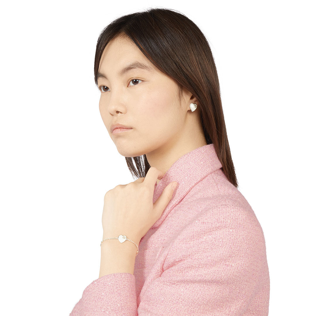 A woman with straight brown hair wears a light pink textured jacket, silver heart-shaped earrings, and the GUCCI Fine Jewellery Gucci Interlocking G Heart Enamel Sterling Silver Bracelet, posing against a plain white background.