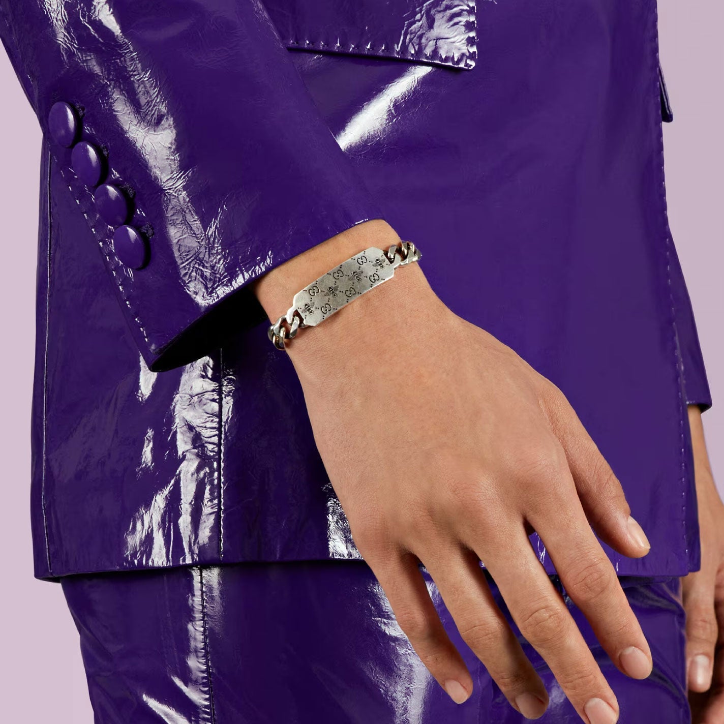 A person wearing a shiny purple suit is shown with their arm extended, showcasing the sparkling Gucci GG and Bee Engraved Sterling Silver Bracelet by GUCCI Fine Jewellery. The sterling silver chain features a rectangular plate engraved with symbols such as hearts, diamonds, and clubs. The background is light purple.