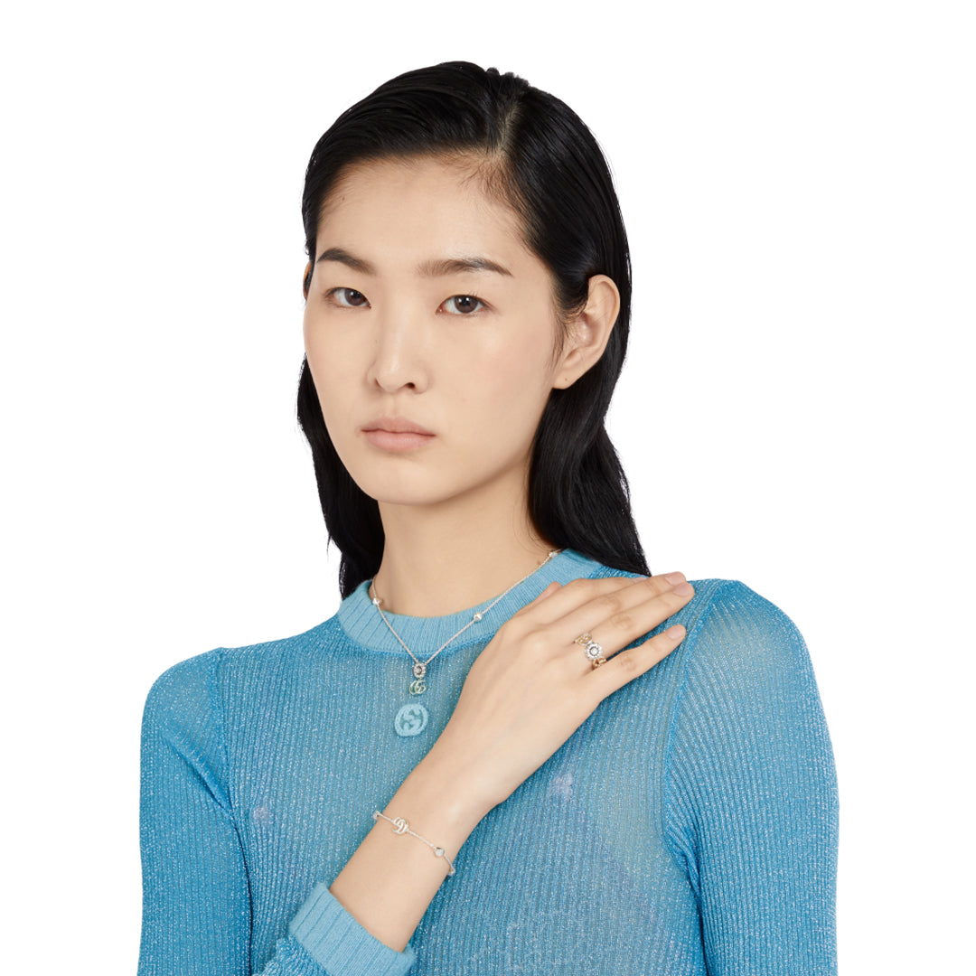 A person with long dark hair wears a sheer blue top and sterling silver jewelry, including a Gucci GG Marmont Sterling Silver Bracelet by GUCCI Fine Jewellery. Against a white background, they gaze directly at the camera with one hand resting on their chest.