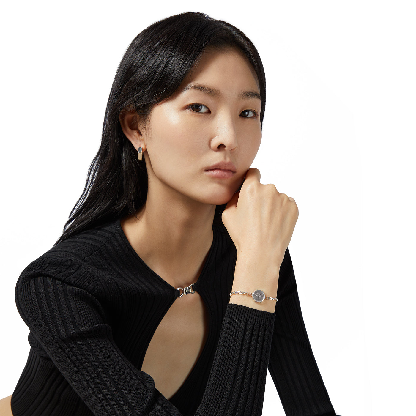 A person with long dark hair, dressed in a black ribbed top, poses with their right hand by their face. They are accessorized with sterling silver earrings and a GUCCI Fine Jewellery Trademark Sterling Silver Bracelet, seated against a plain white backdrop.