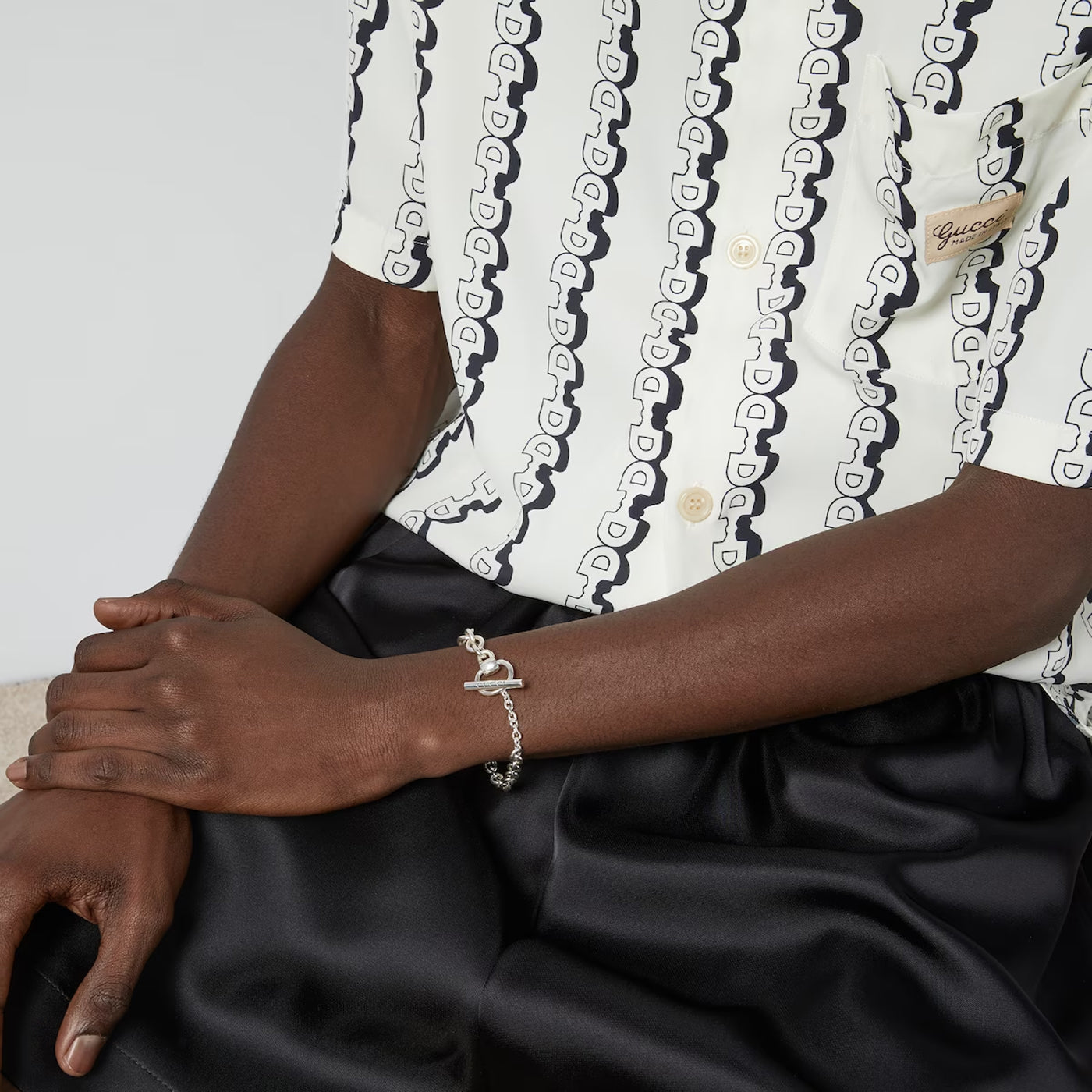 A person wearing a shirt with interlocking patterns and black pants sits with their hands gently crossed. They wear a Gucci Horsebit Chain Sterling Silver Bracelet featuring a snake design, crafted by GUCCI Fine Jewellery. The shirt, featuring an equestrian-inspired toggle closure, has a chest pocket with a subtle logo.