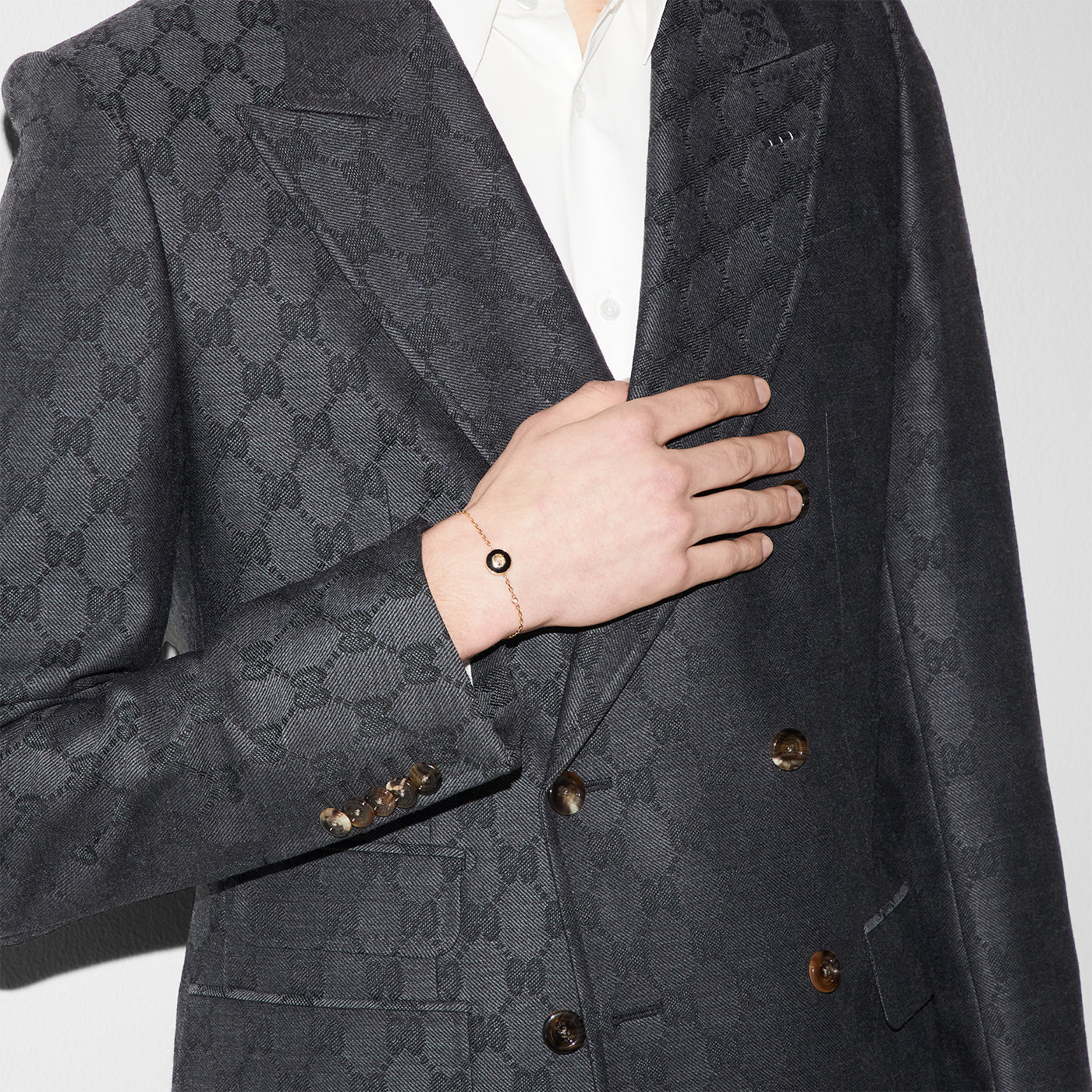 A person in a gray, patterned blazer and white shirt poses with a hand placed on the chest, showcasing the elegant Gucci Interlocking G 18K Yellow Gold Chain Bracelet With Onyx. The blazer features dual buttons and a subtle design, while the Interlocking G detail from GUCCI Fine Jewellery adds sophistication to the ensemble against the plain white background.