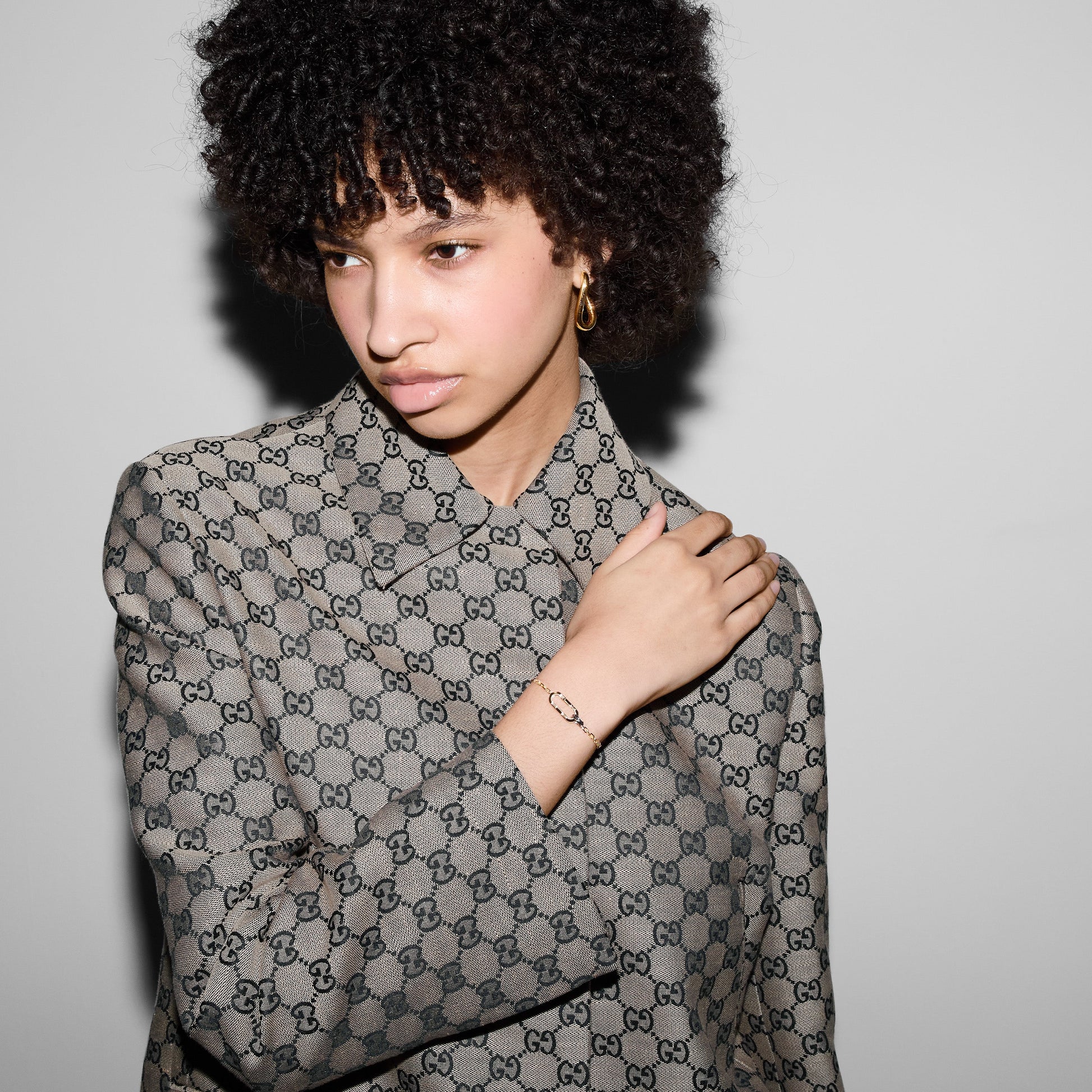 A person with curly hair, dressed in a patterned jacket and adorned with 18k yellow gold hoop earrings, glances to the side while touching their shoulder. The plain, light-colored background accentuates their fashion ensemble, subtly evoking the style of the GUCCI Fine Jewellery's Link To Love collection.