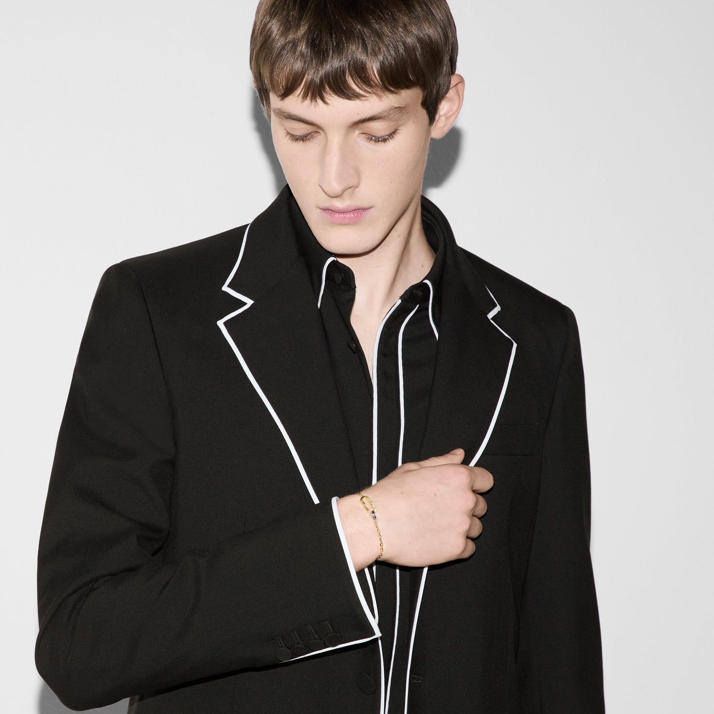 A person with short brown hair is dressed in a black suit jacket featuring white piping, layered over a black shirt. They are gazing downward, with their left hand placed on their chest where a GUCCI Fine Jewellery 18K Yellow Gold Link To Love Chain Bracelet adorns their wrist. The background is plain white.