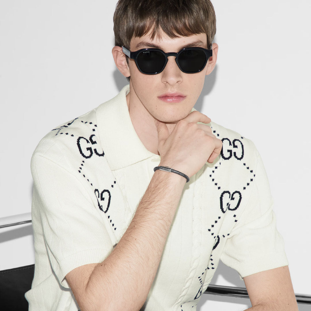 A person with short brown hair is wearing black sunglasses and a light-colored polo shirt featuring black geometric patterns. Their wrist is adorned with a GUCCI Fine Jewellery trademark GG Canvas with Sterling Silver Bracelet as they sit against a plain white background, resting their chin on their hand.