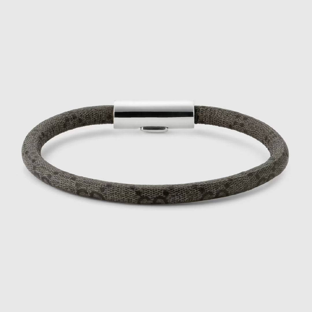 The Gucci Trademark GG Canvas with Sterling Silver Bracelet from GUCCI Fine Jewellery boasts a stylish design, featuring a flexible dark band that might be made from GG Supreme canvas, complemented by a shiny metallic cylindrical sterling silver clasp. Its modern and minimalist aesthetic is beautifully contrasted against a light gray background.