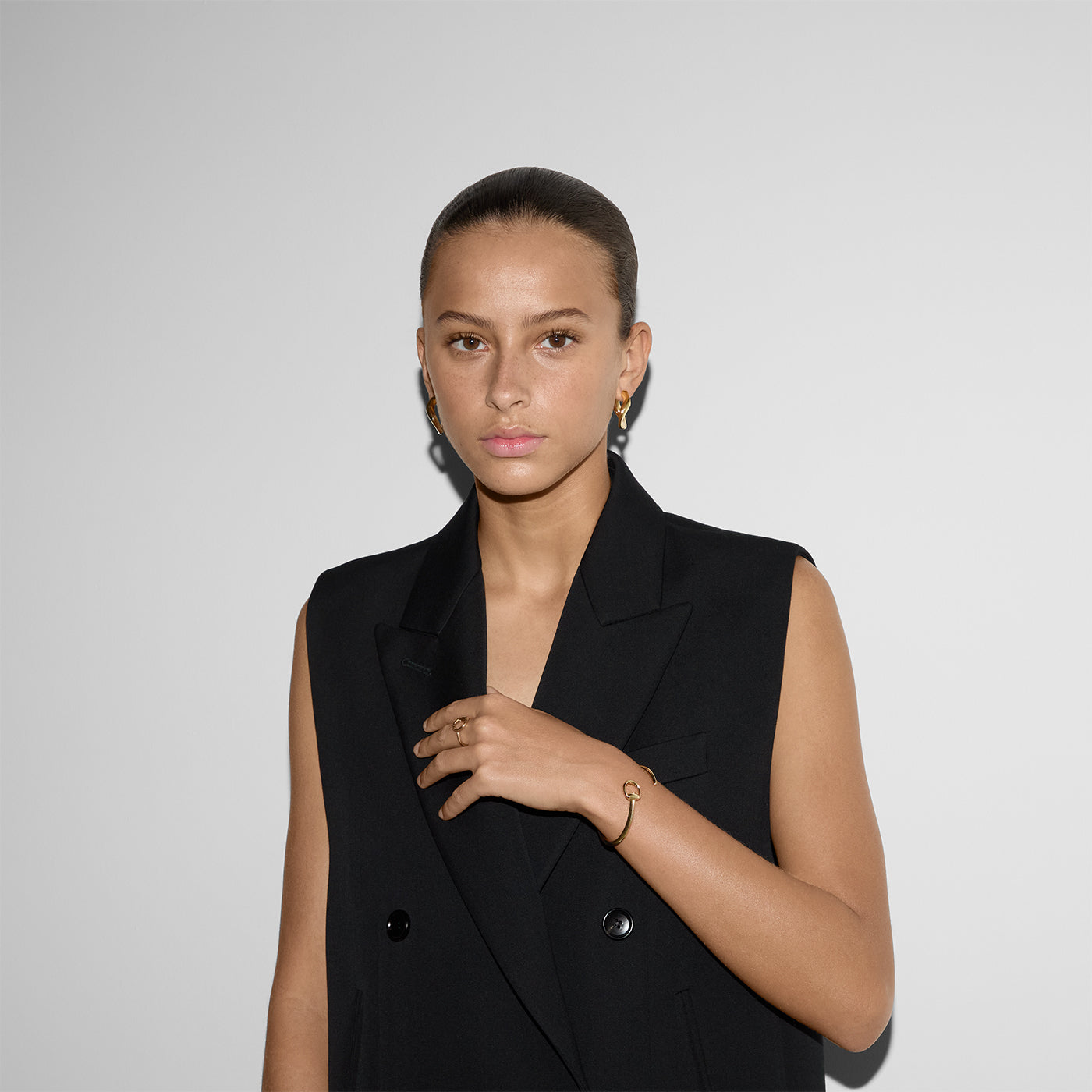 A woman with tied-back hair wears a sleeveless black blazer and stands against a white backdrop, looking directly at the camera. She accessorizes with small hoop earrings, a ring, and a signature piece—a Gucci Horsebit 18K Yellow Gold Cuff Bracelet by GUCCI Fine Jewellery.