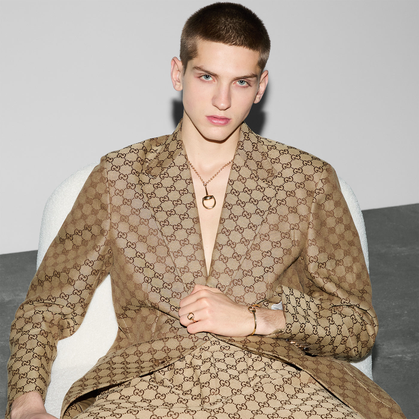 A person with short hair sits in a chair, dressed in a patterned brown suit featuring a distinctive signature detail. They are adorned with the Gucci Horsebit 18K Yellow Gold Cuff Bracelet from GUCCI Fine Jewellery and gaze thoughtfully. The setting is minimalistic, showcasing a two-tone wall and a gray floor.