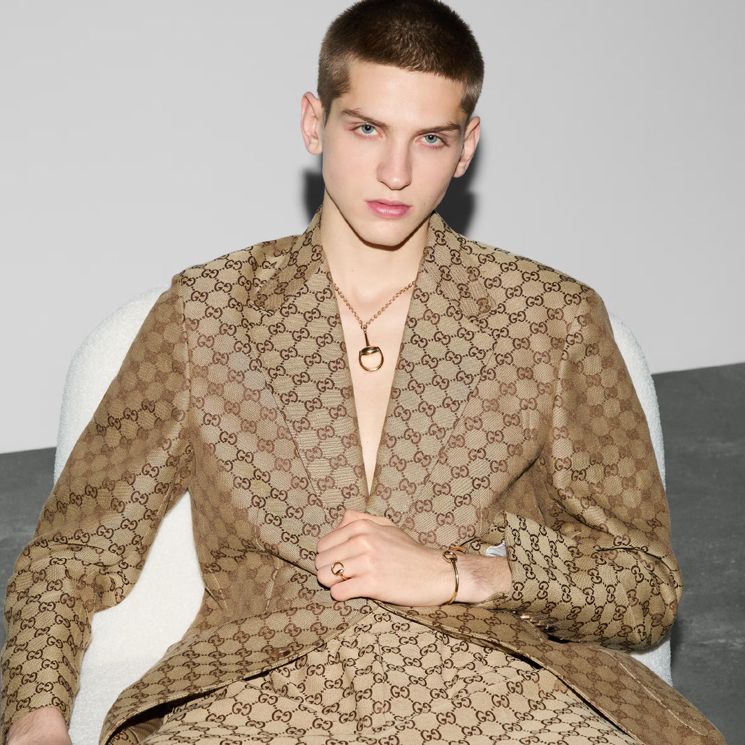 A person with short hair sits on a white chair, wearing a tan Gucci suit with logo pattern, accessorized with the GUCCI Fine Jewellery Horsebit 18K Yellow Gold Cuff. The background is minimal and gray.
