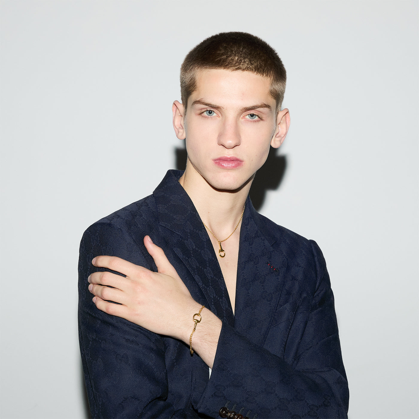 A person with short hair stands against a simple backdrop, clad in a dark patterned blazer. They accessorize with GUCCI Fine Jewellery, including a Gucci Horsebit 18K yellow gold necklace and the Gucci Horsebit 18K Yellow Gold Chain Bracelet, their hand gently resting on their opposite shoulder.