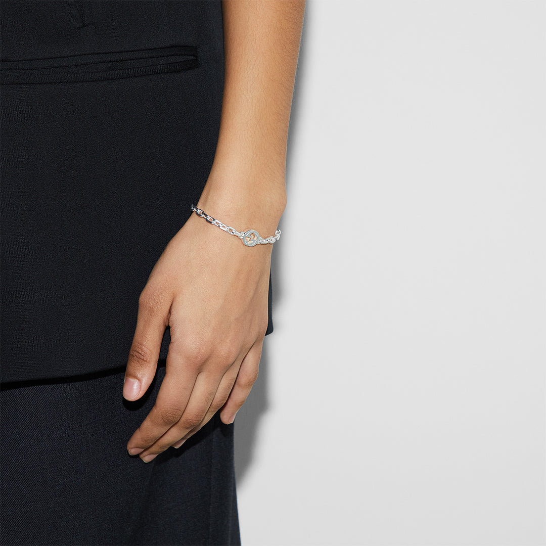 Dressed in black, a person displays the Gucci Interlocking G Sterling Silver Bracelet by GUCCI Fine Jewellery, featuring an infinity symbol and elegant Interlocking G motif against a plain white wall.