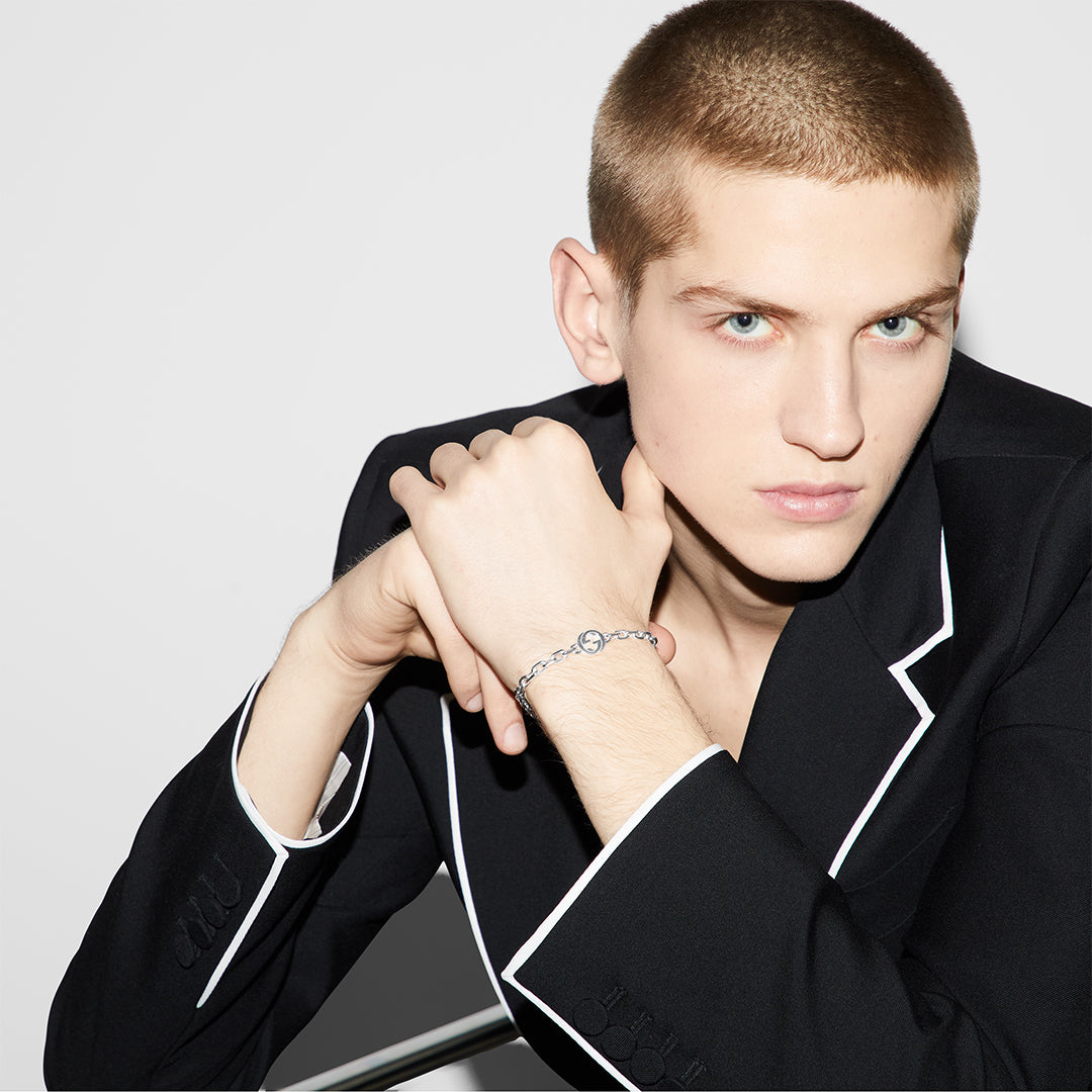 Dressed in a black jacket with white trim, a person with short hair rests their chin on their hand, highlighting the Gucci Interlocking G Sterling Silver Bracelet by GUCCI Fine Jewellery. Set against a plain white background, their style exudes sophistication and elegance.