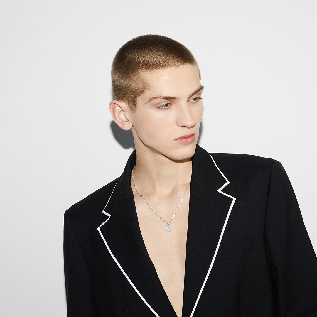 A person with short hair wearing a black blazer with white piping, gazing to the side against a plain white background, reveals an elegant Gucci Interlocking G Sterling Silver Necklace Pendant by GUCCI Fine Jewellery that subtly complements the open-chest ensemble.