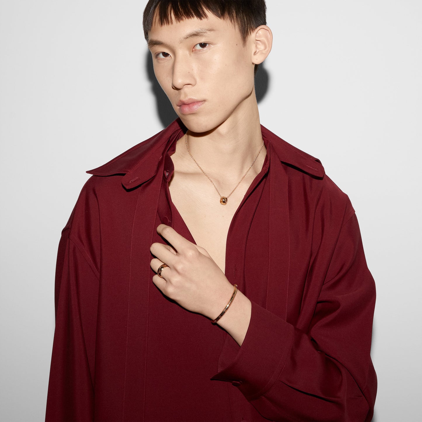 A person dressed in a maroon shirt stands out against a white backdrop, with short hair and an array of stunning accessories. They sport the Gucci Icon 18K Yellow Gold GG Bangle from GUCCI Fine Jewellery, paired with a matching GG cut-out motif necklace and ring. Elegantly grabbing their shirt collar with one hand, their ensemble perfectly showcases Gucci's luxurious style.