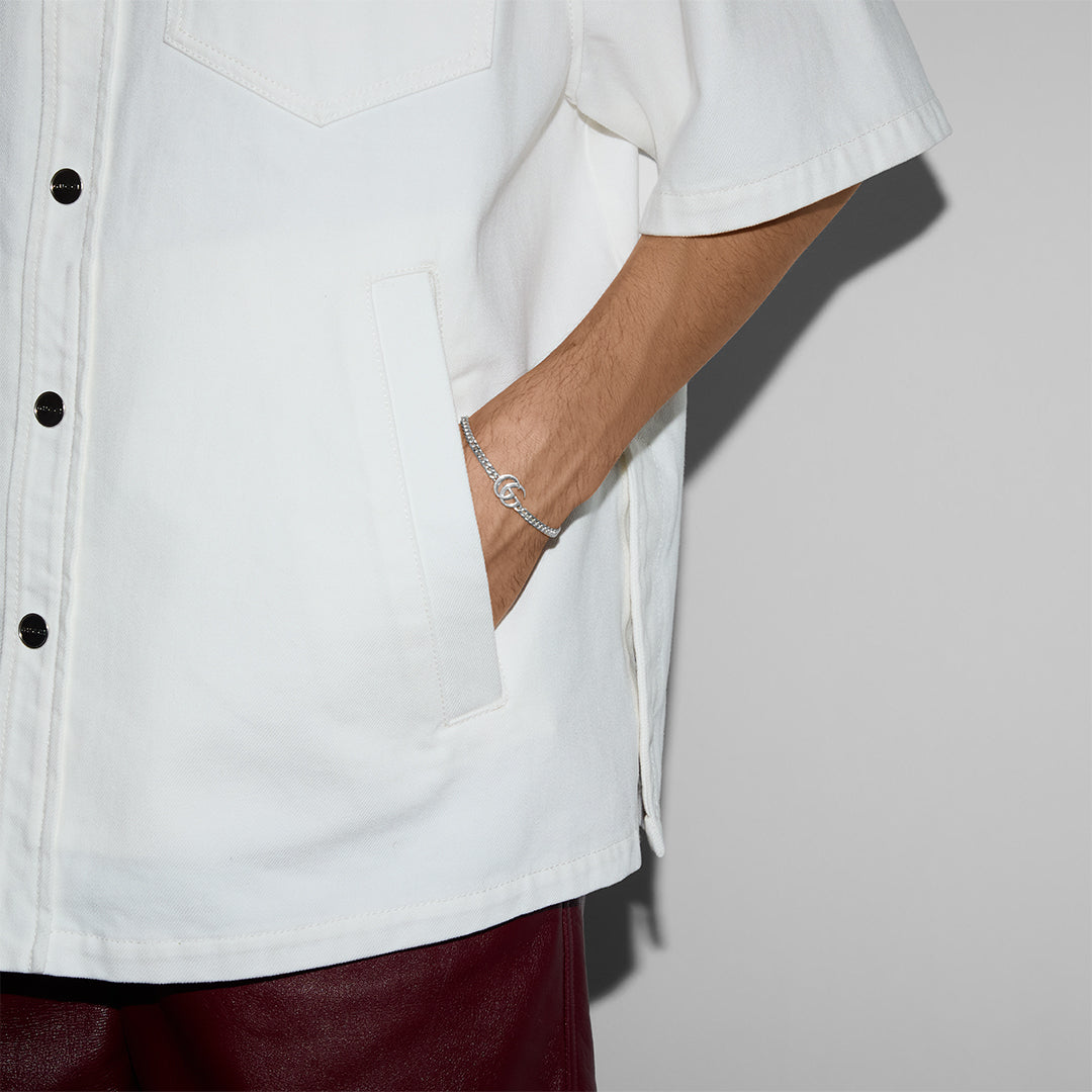 A person wears a white short-sleeve button-up shirt with black buttons, dark red pants, and their right hand in a pocket, accessorized with a GUCCI Fine Jewellery Gucci GG Marmont Sterling Silver Chain Bracelet. The background is a plain light gray wall.