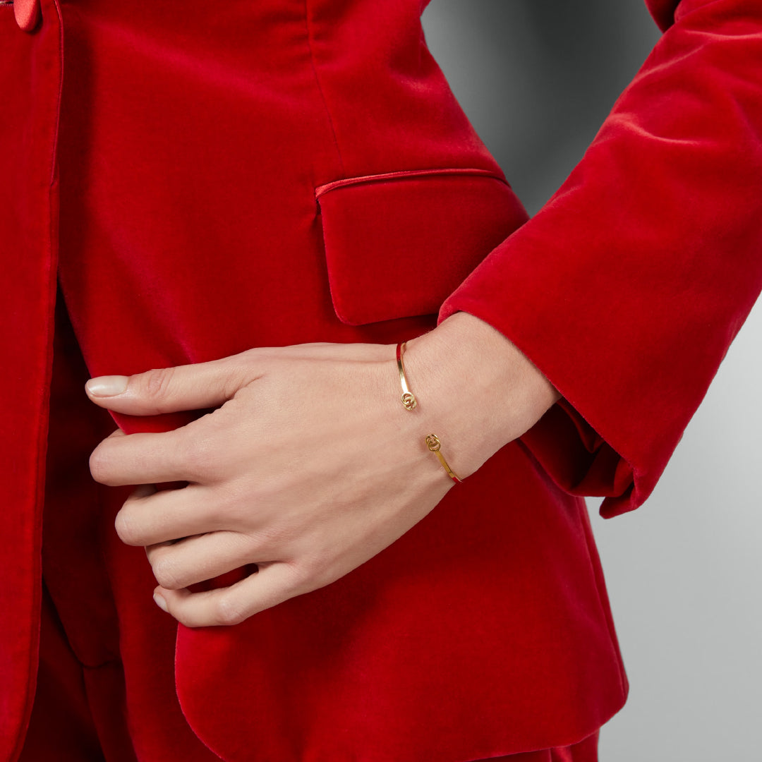 A person in a vibrant red velvet blazer rests their hand near the pocket, displaying the GUCCI Fine Jewellery Gucci GG Running 18K Yellow Gold Cuff on their wrist against a neutral background.