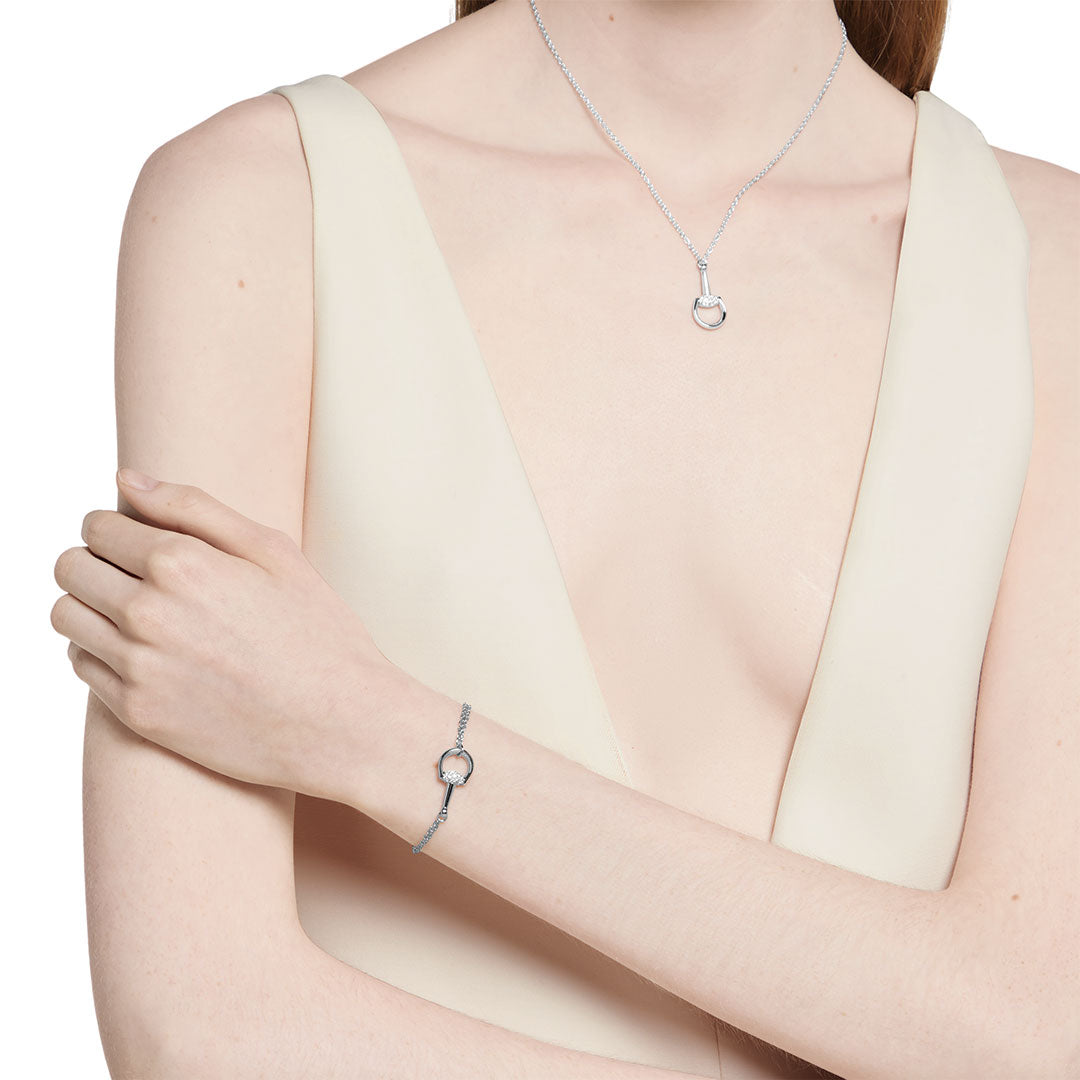 A woman in a sleeveless ivory dress wears the Gucci Horsebit 18K White Gold Diamond Bracelet by GUCCI Fine Jewellery and a matching necklace with circular pendants. Her arms are crossed, and her long hair falls over one shoulder.