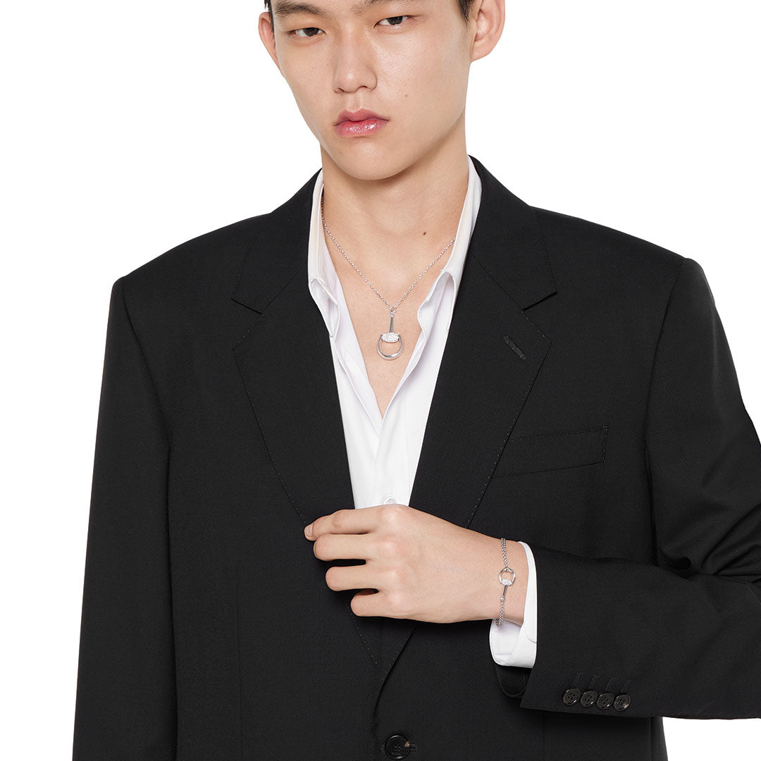 A person in a black suit jacket and white shirt, accessorized with a GUCCI Fine Jewellery Horsebit 18K White Gold Diamond Bracelet and a matching silver necklace, looks forward with a neutral expression.