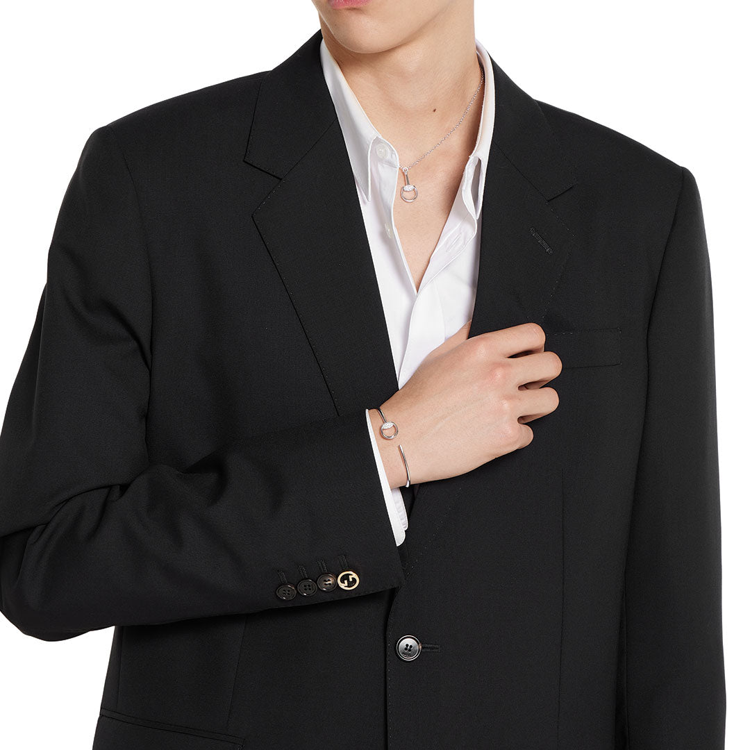 Dressed in a black suit and white shirt, a person elegantly displays the GUCCI Fine Jewellery Horsebit 18K White Gold Diamond Cuff Bracelet by raising their hand to their chest.