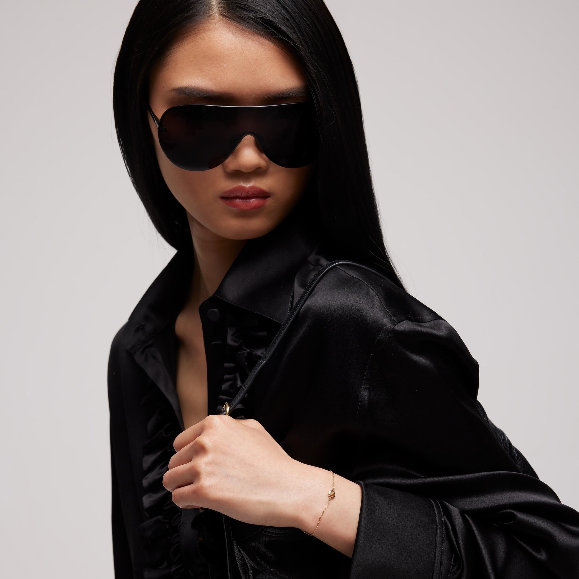 A woman in large black sunglasses and a black satin shirt looks to the side, holding her collar. On her wrist is the Gucci Interlocking 18K Yellow Gold Chain Bracelet by GUCCI Fine Jewellery. The background is plain and light-colored.