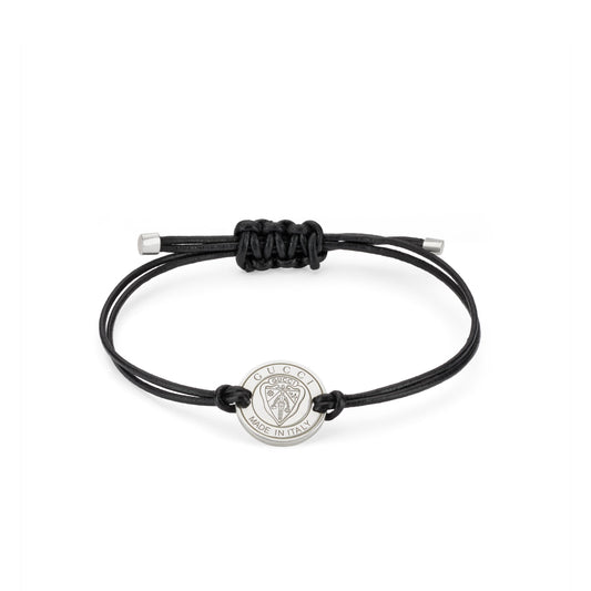 The Gucci Crest Leather Cord with Sterling Silver Bracelet by GUCCI Fine Jewellery features a minimalist black leather cord, an adjustable knot, and a sterling silver charm engraved with "GUCCI" and "MADE IN ITALY," showcasing the iconic lion head crest.