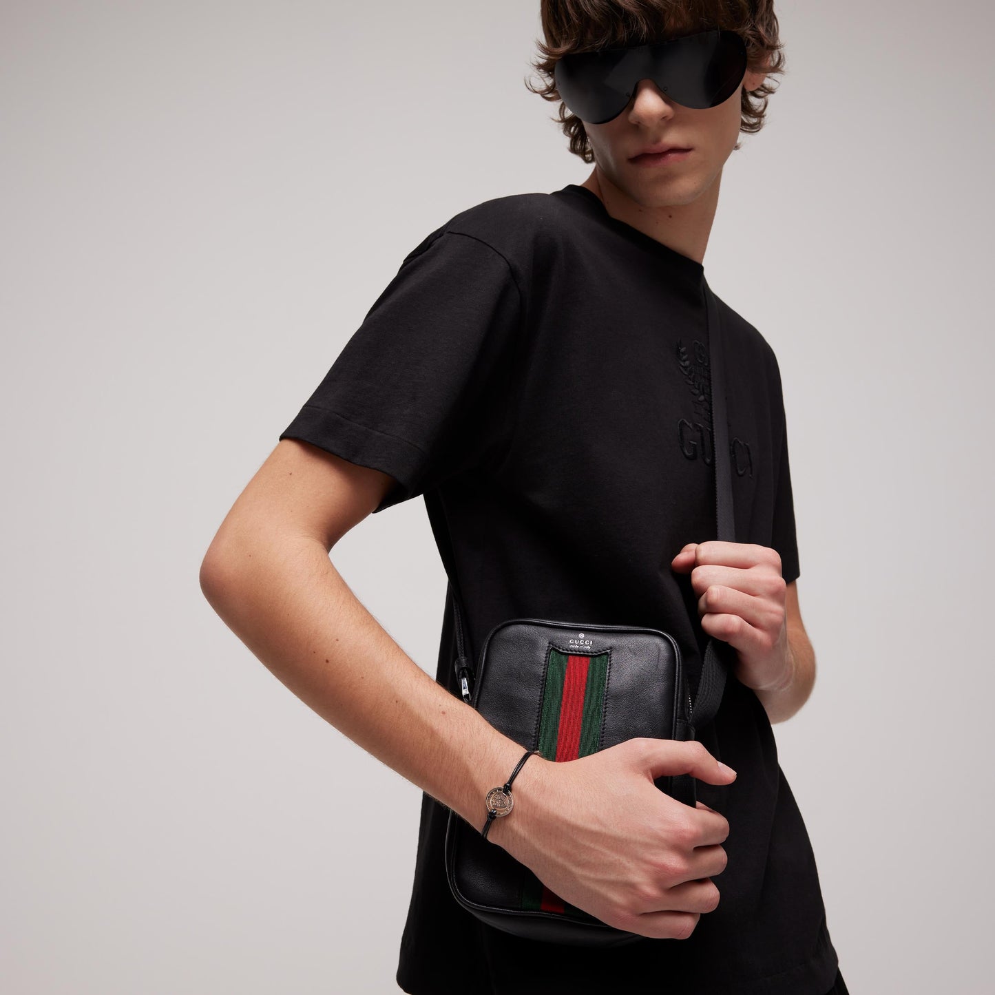 A young person poses against a plain background wearing black sunglasses and a black t-shirt, showcasing the Gucci Crest Leather Cord with Sterling Silver Bracelet from GUCCI Fine Jewellery.