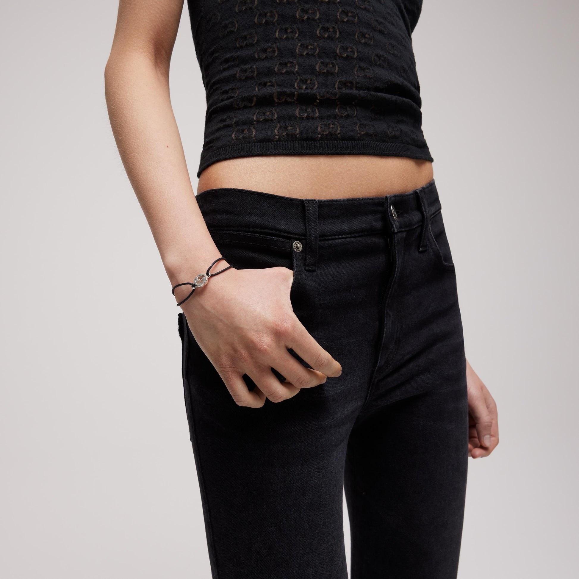 A person in a black sleeveless patterned top and high-waisted black jeans stands with one hand down, displaying the GUCCI Fine Jewellery Interlocking Cotton Cord with Sterling Silver Bracelet on their wrist.