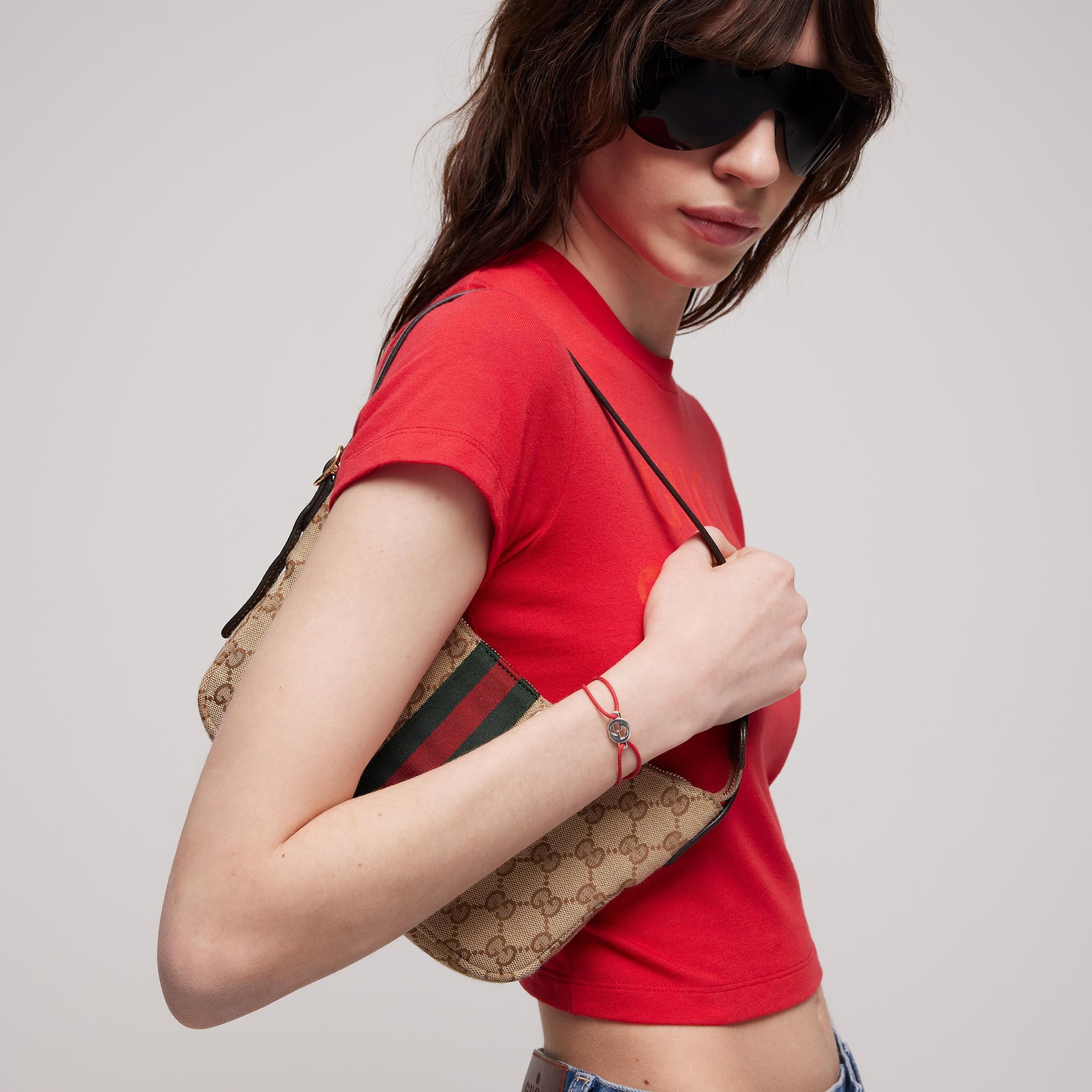 A person in a red crop top, sunglasses, and jeans holds a beige Gucci handbag with green and red stripes, accessorized with the GUCCI Fine Jewellery Interlocking Cotton Cord with Sterling Silver Bracelet, against a plain background.