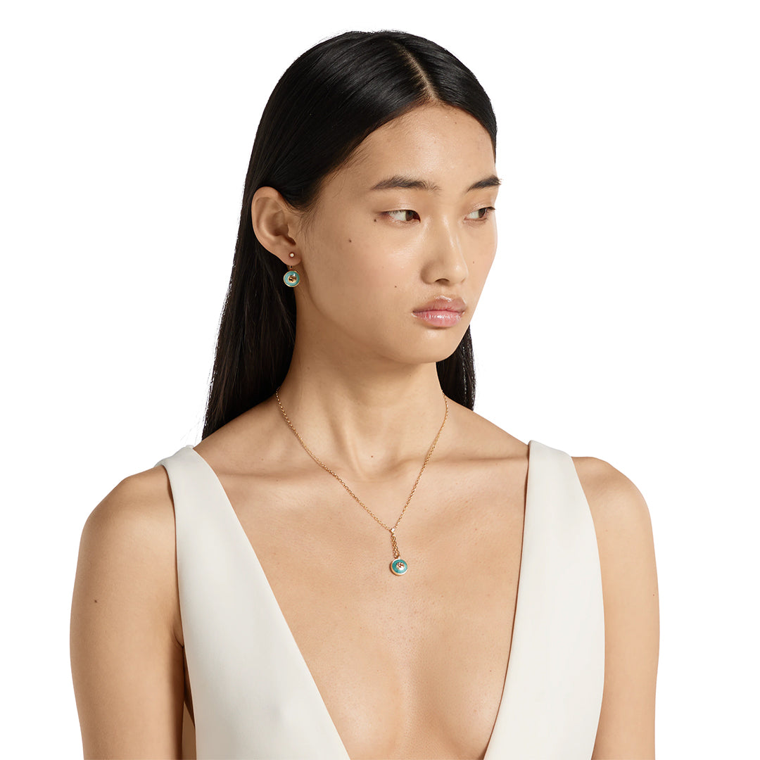 A woman with long black hair, in a sleeveless deep v-neck white top, wears the GUCCI Fine Jewellery Interlocking 18K Yellow Gold Diamond Necklace Pendant With Amazonite and matching earrings, looking slightly left against a plain white background.
