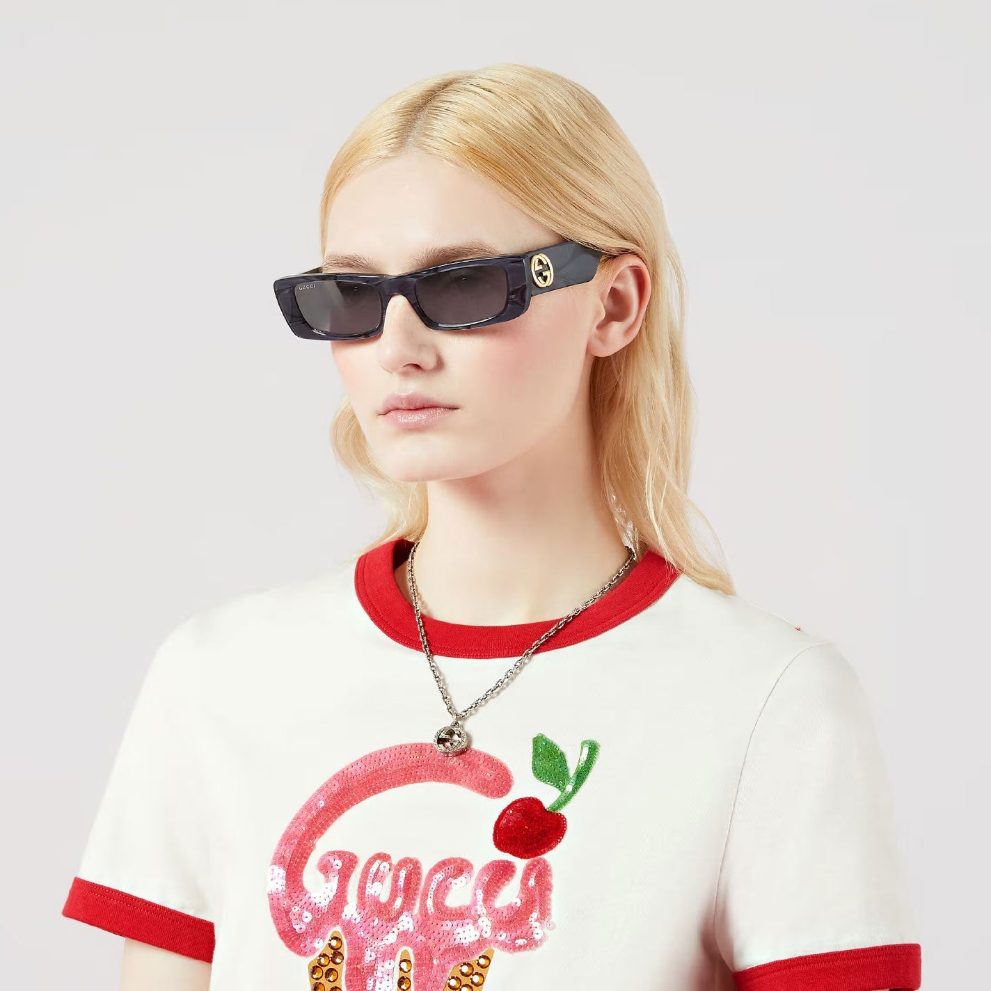 A person with long blonde hair is wearing black rectangular sunglasses and a white T-shirt featuring a cherry design. They are accessorized with the Gucci Interlocking G Sterling Silver Necklace from GUCCI Fine Jewellery, standing against a plain background.