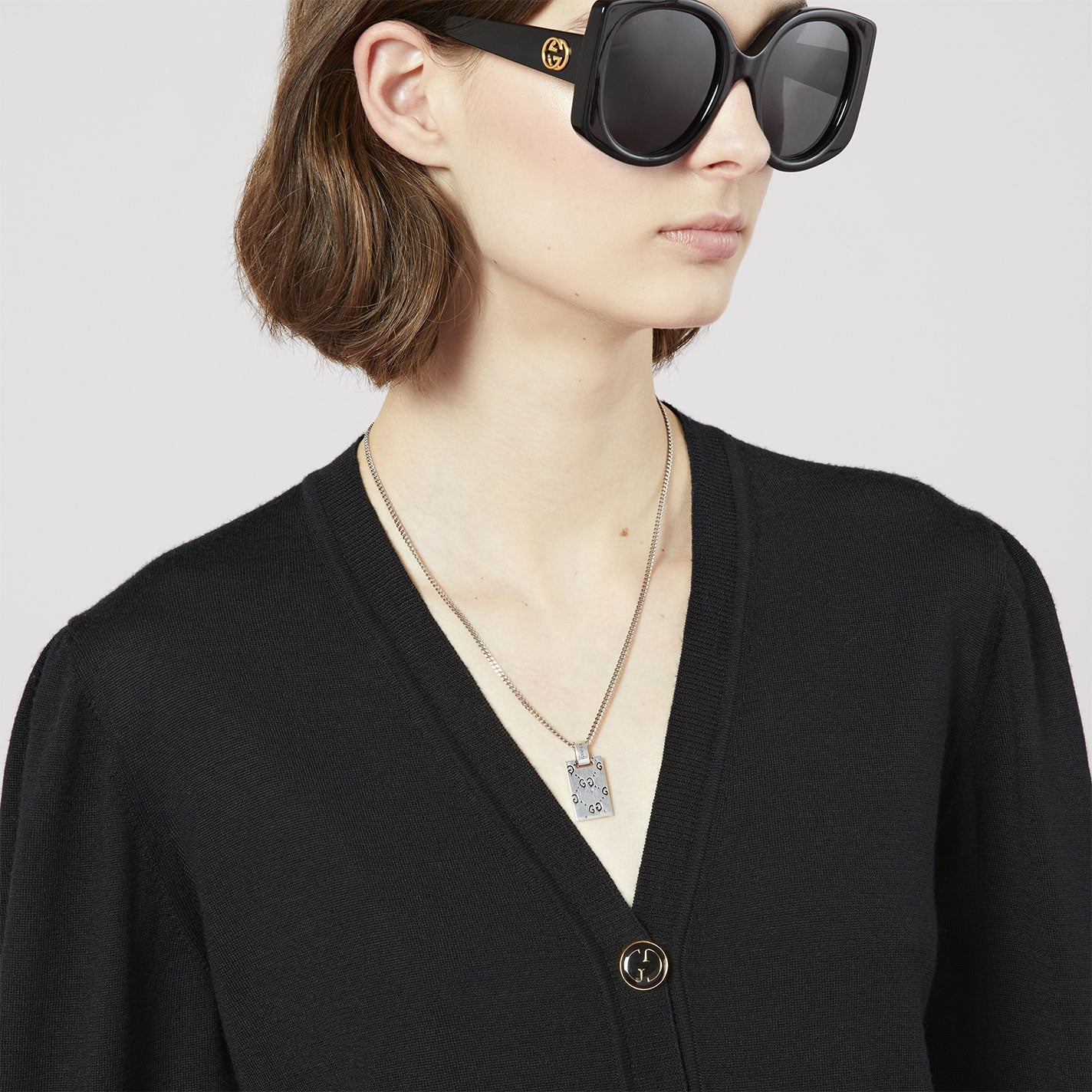 A person with short brown hair dons oversized black sunglasses and a black V-neck sweater adorned with a gold button. They are accessorized with a layered sterling silver necklace featuring the Gucci Ghost Sterling Silver Necklace Pendant from GUCCI Fine Jewellery. The backdrop is plain and light-colored.