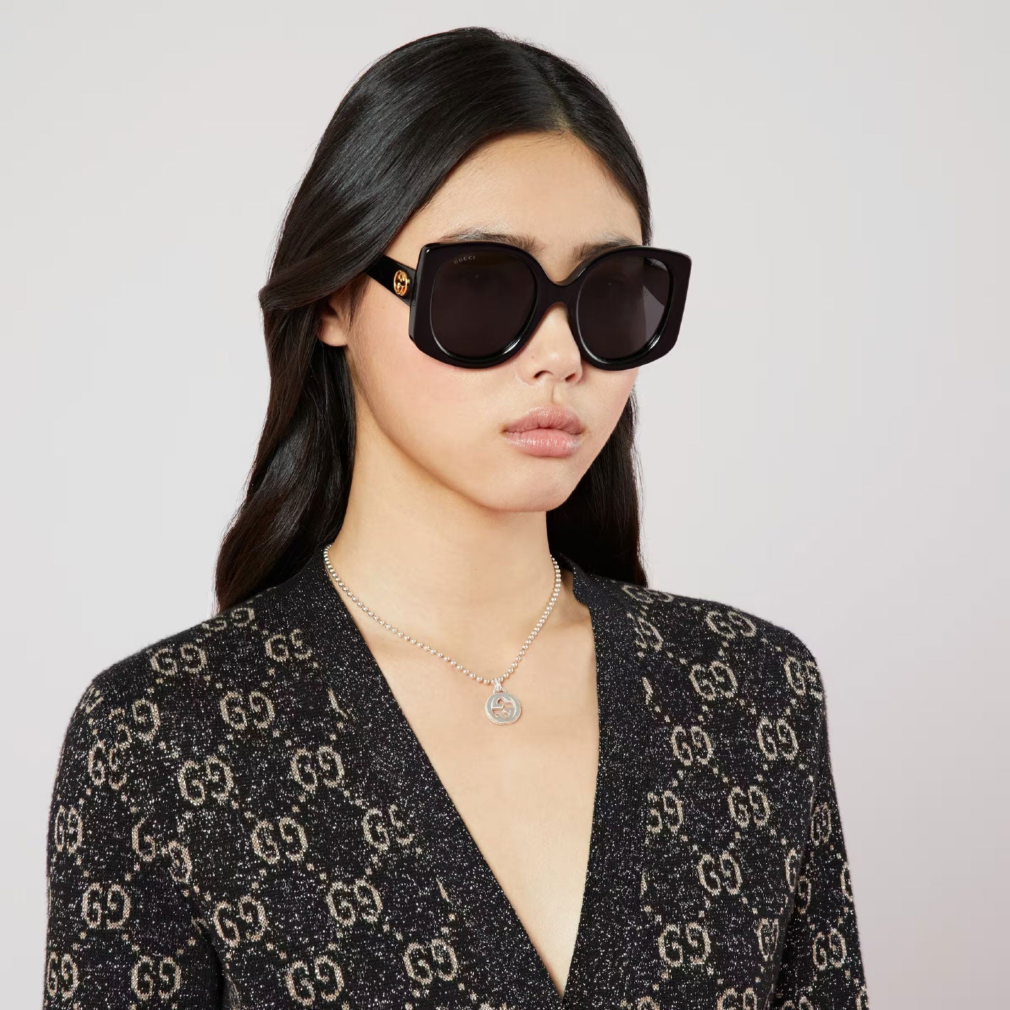 A woman with long dark hair dons oversized black sunglasses and a patterned black top. Her expression is neutral, and she is accessorized with a striking GUCCI Fine Jewellery pendant necklace featuring the iconic Interlocking G design, crafted in sterling silver, set against a plain background.