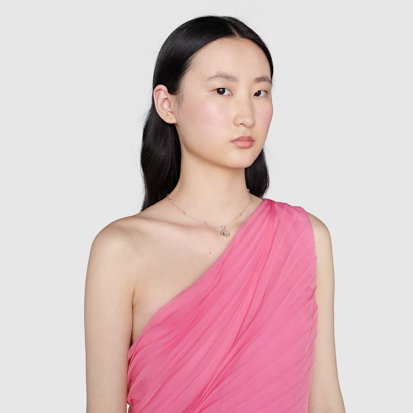 A person with long black hair wears a pleated pink one-shoulder dress and a delicate Gucci Double G Pink Mother of Pearl Sterling Silver Station Necklace Pendant by GUCCI Fine Jewellery. They have a neutral expression and stand against a plain gray background.