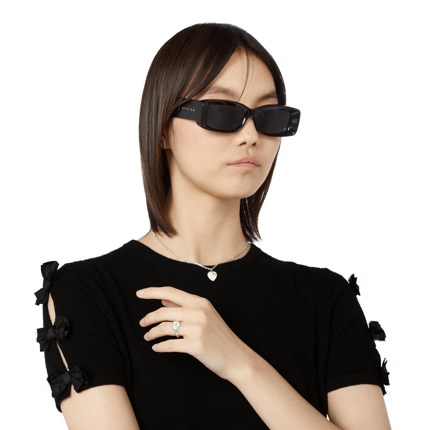 A person with straight brown hair dons rectangular black sunglasses and a black top with bows on the sleeves. They wear a GUCCI Fine Jewellery necklace featuring the Interlocking G Sterling Silver Heart Enamel Pendant, and showcase a ring against a plain white background, adding elegance.