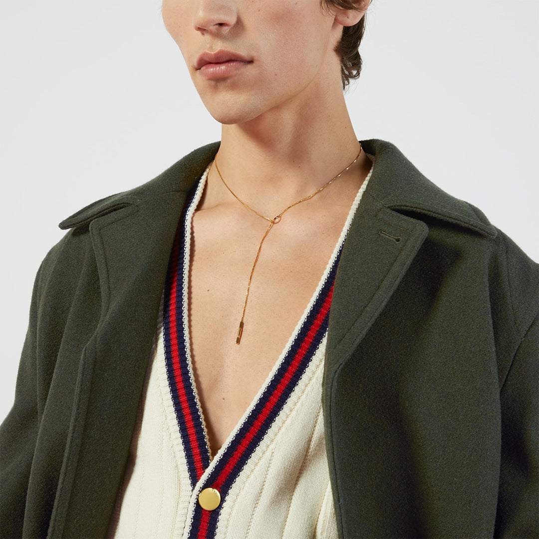 A person wears a dark green coat over a cream deep V-neck cardigan with red and navy stripes, accessorized with a GUCCI Fine Jewellery Link To Love 18K Yellow Gold Lariat Necklace. Only the lower face and upper torso are visible.