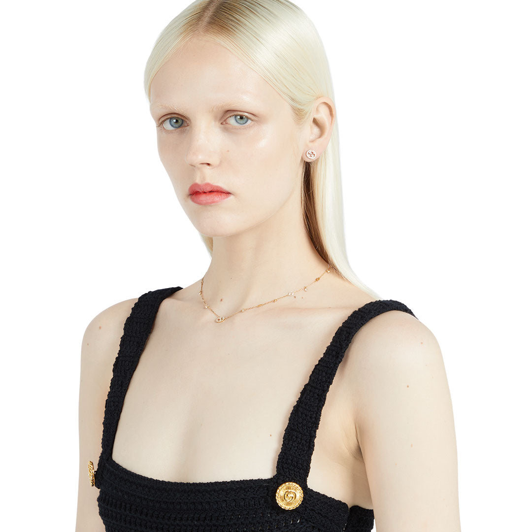 A woman with long, straight platinum blonde hair wears a black textured sleeveless top with gold buttons and small earrings. She accessorizes with the GUCCI Fine Jewellery Interlocking G 18K Yellow Gold Diamond Station Necklace Pendant.