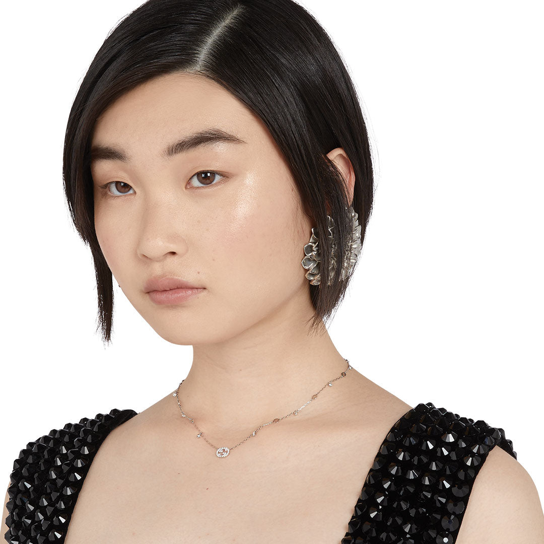 A woman with a short black bob wears a textured black top, bold silver earrings, and the GUCCI Fine Jewellery Interlocking G 18K White Gold Diamond Station Necklace Pendant. She gazes slightly to the side with a neutral expression.