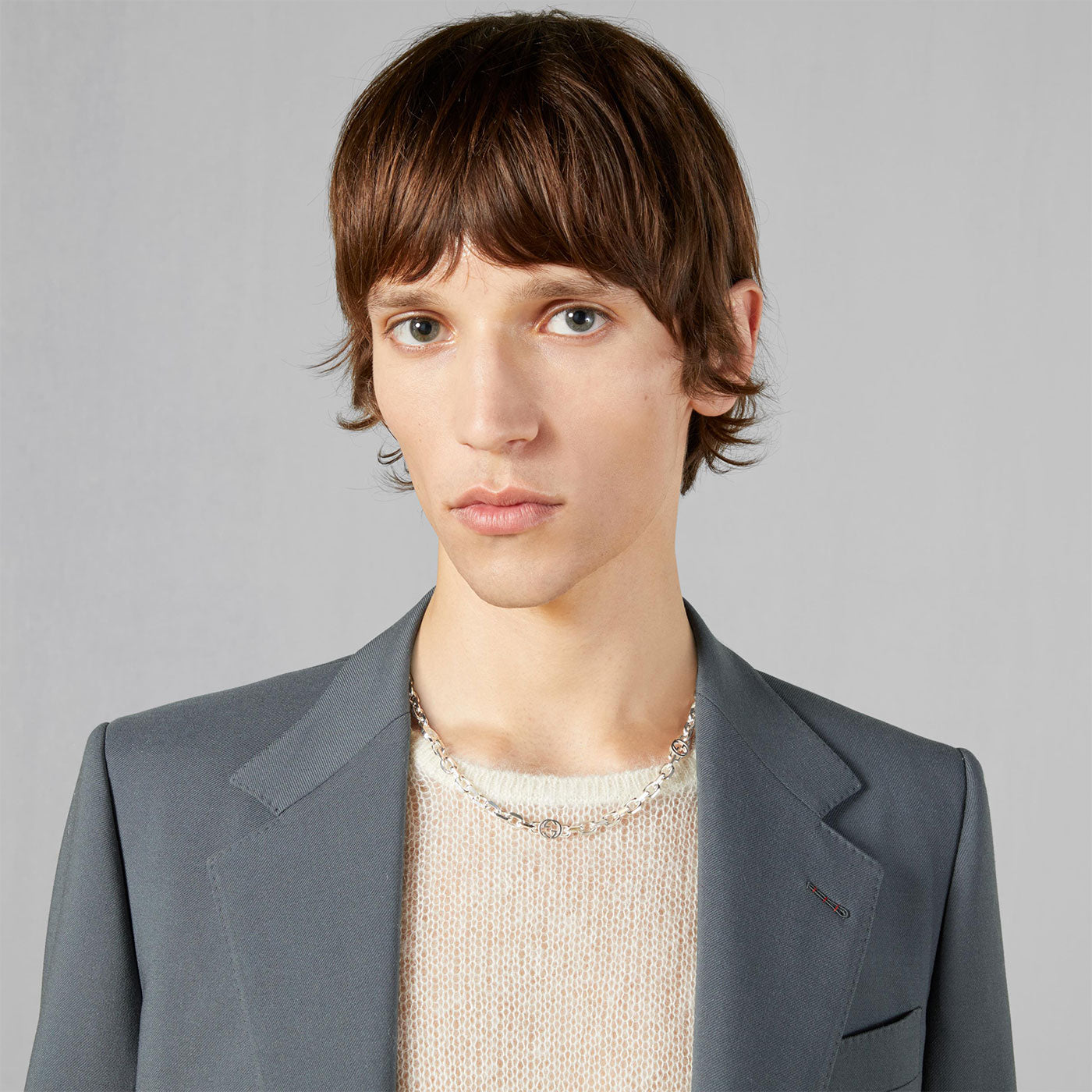 A person with brown hair wearing a grey blazer, a beige sweater, and a Gucci Interlocking G Sterling Silver Chain Station Necklace by GUCCI Fine Jewellery is posing against a plain grey background.